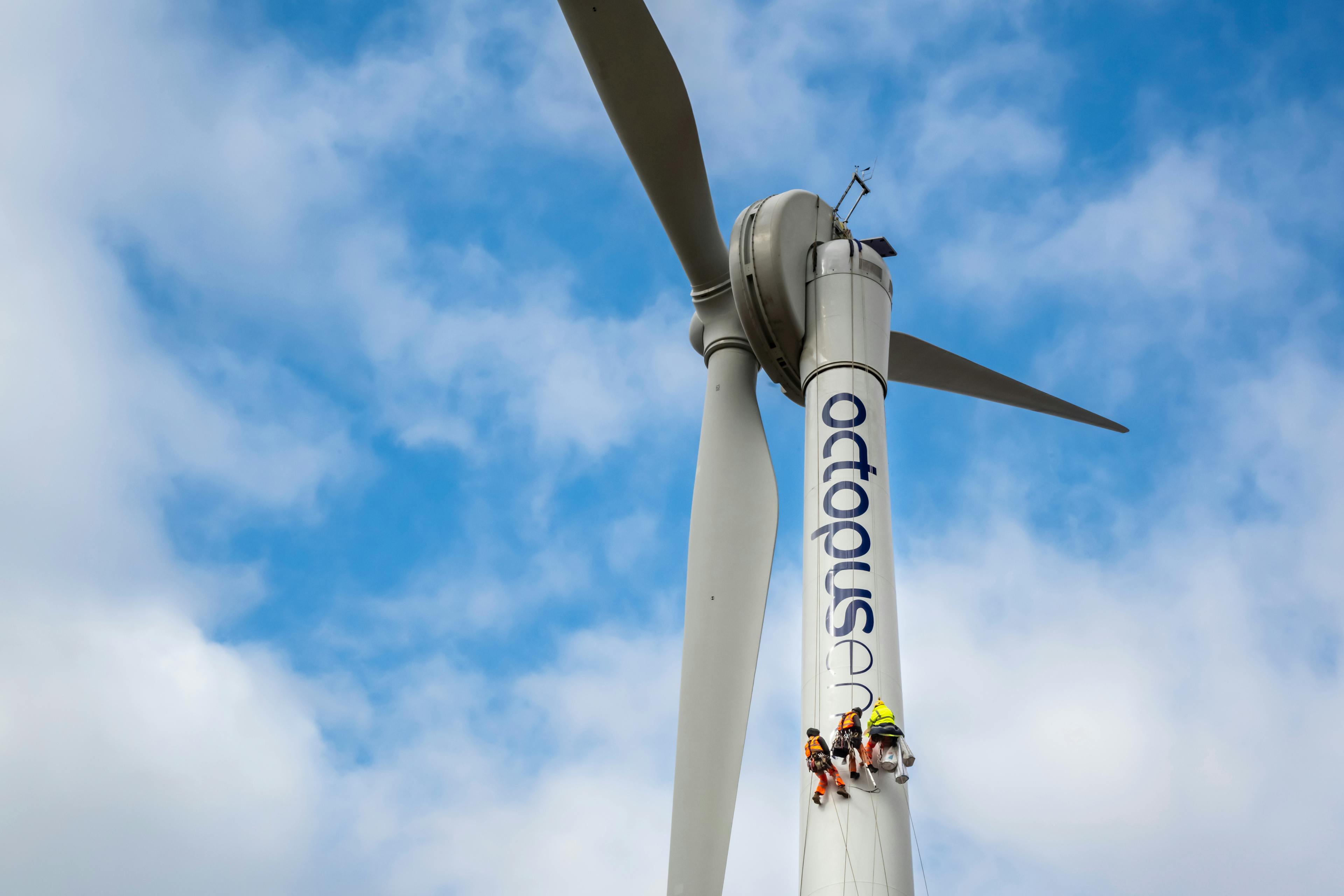 A close-up of a wind turbine in the sky with workers applying the Octopus Energy logo to the stem